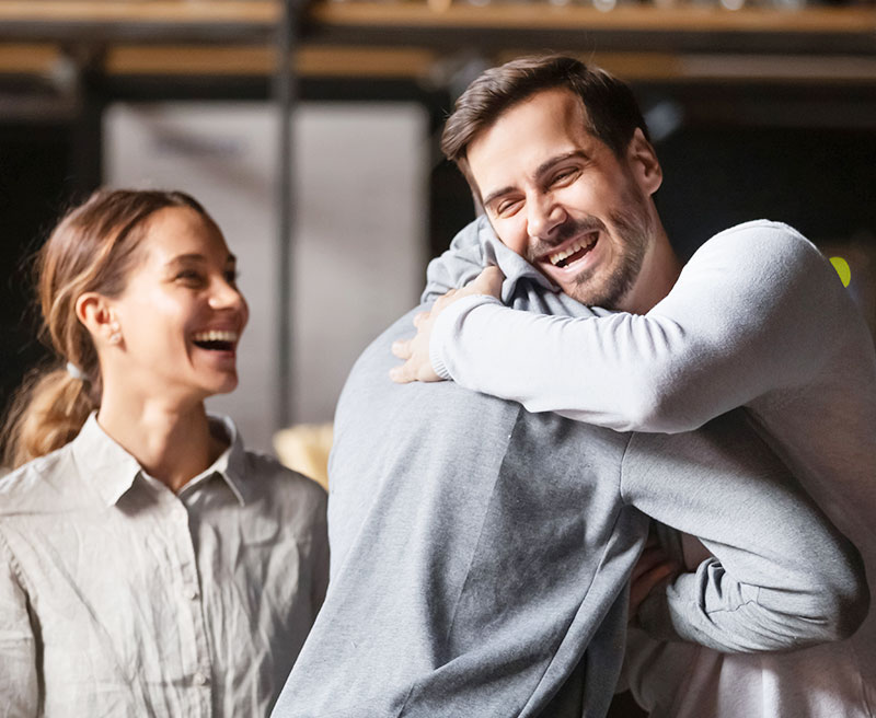 A guy giving a guy a huge with a girl in the background smiling